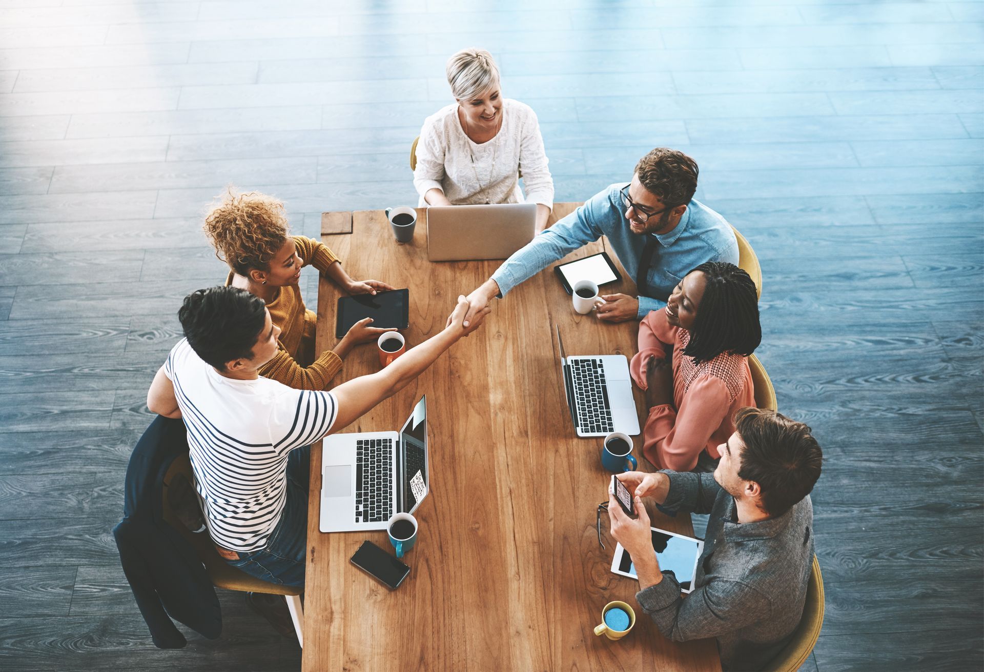 An overhead view of a diverse group shaking hands at a wooden conference table with laptops and documents.