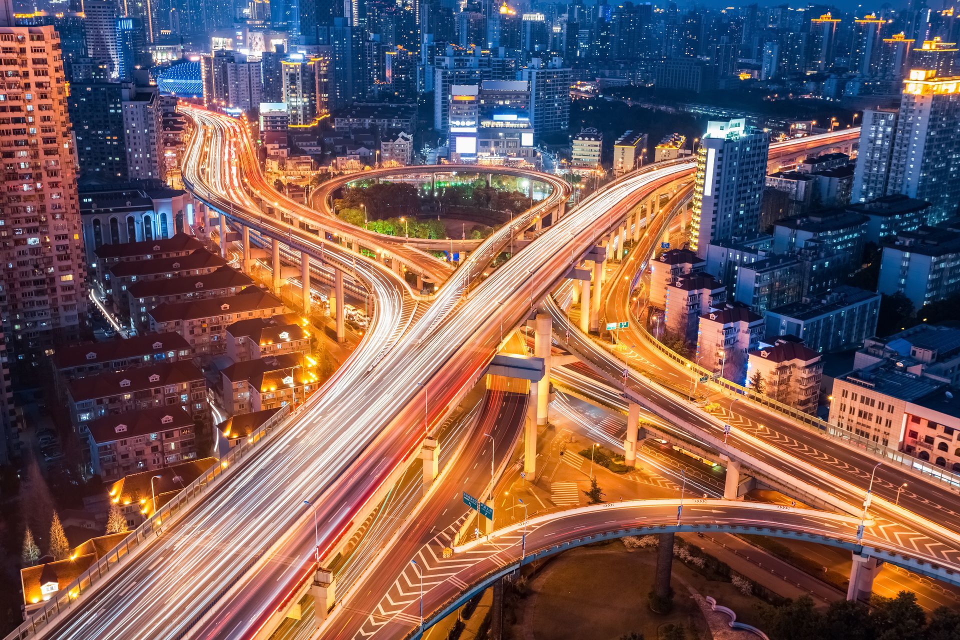 An aerial night view of a sprawling multi-level highway interchange in a city, illuminated by glowing traffic light trails.