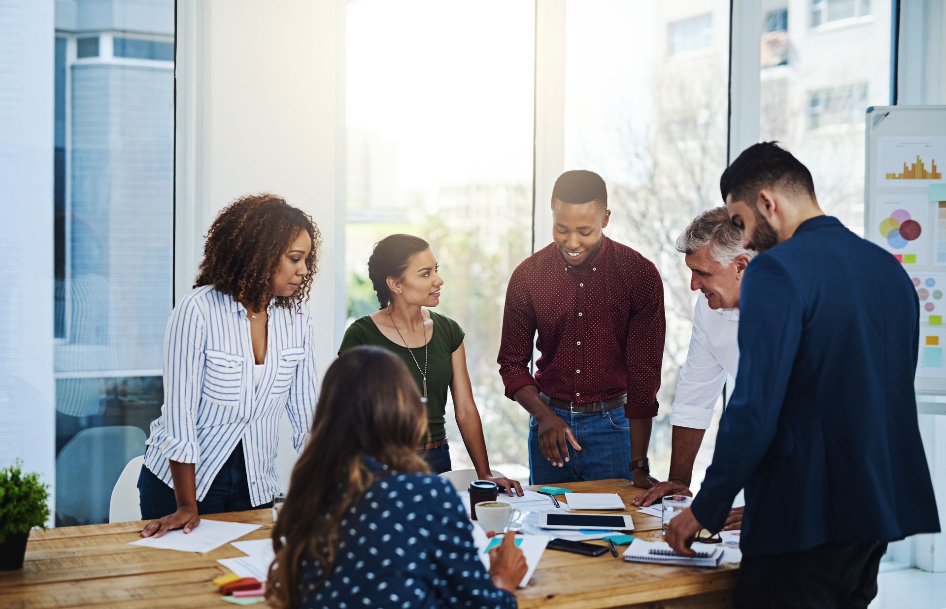 Team collaborates around a table in a bright office, discussing papers, smiling, with city views.