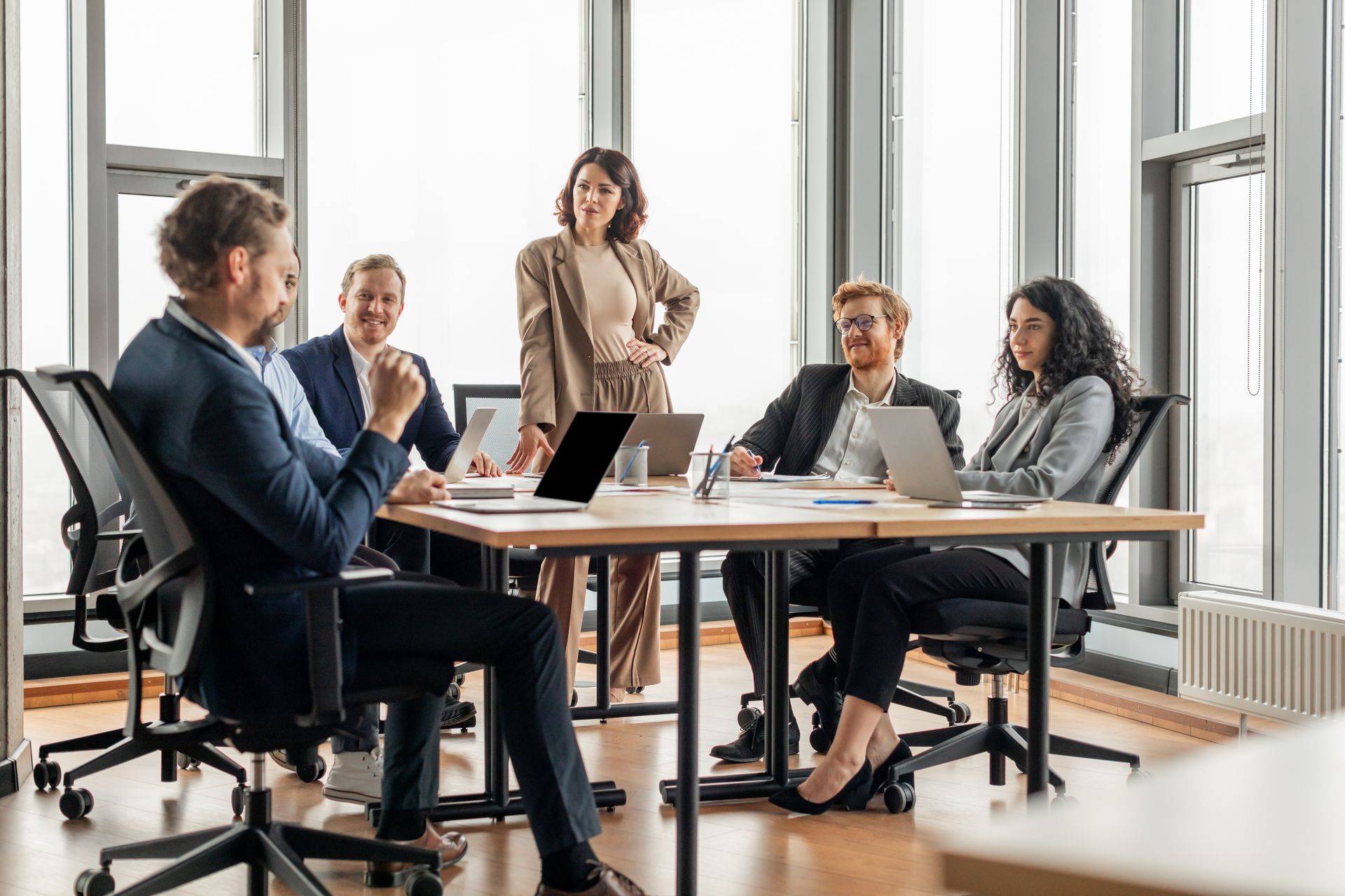 Business team in suits collaborating around a table, one woman standing, laptops present.