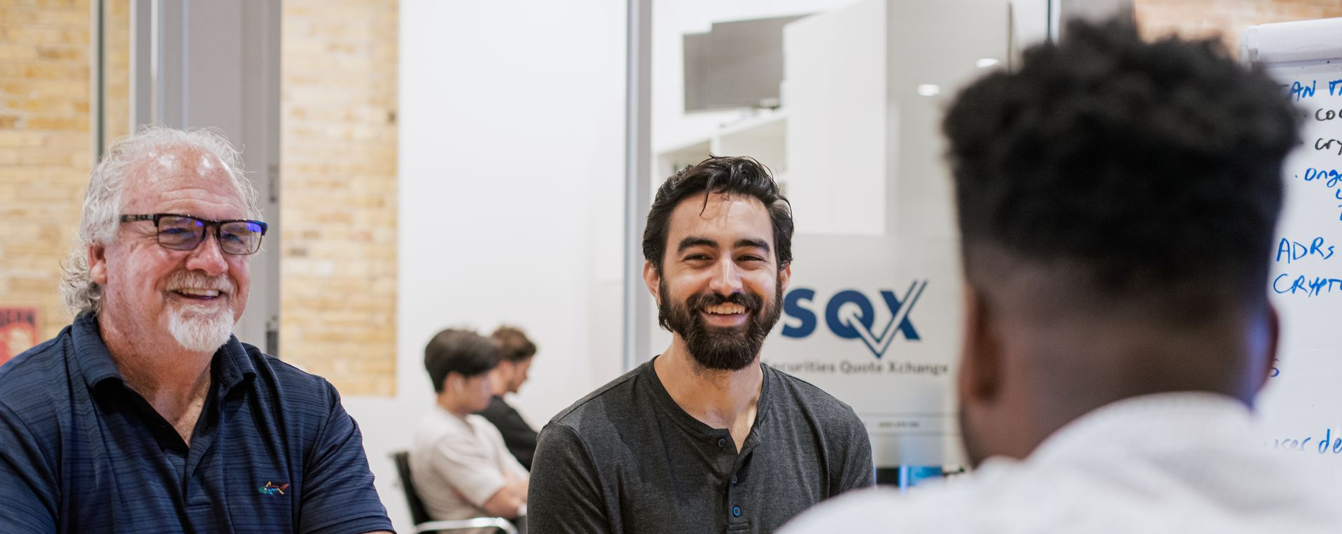 Three men in a meeting, one with a beard and smiling. Another man has his back to the camera.