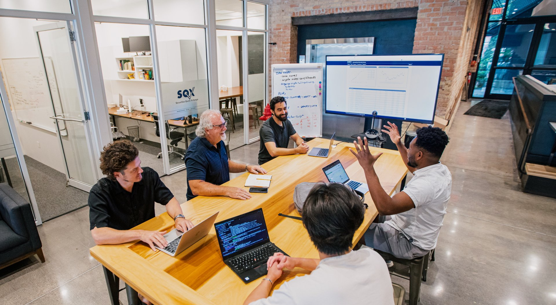 Group of people in an office meeting. They are at a table looking at a screen.