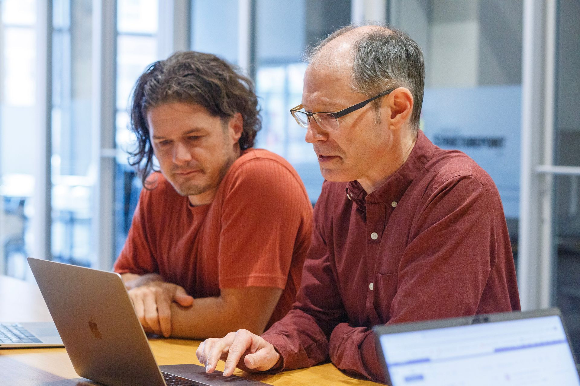 Two men are sitting at a table looking at a laptop computer.