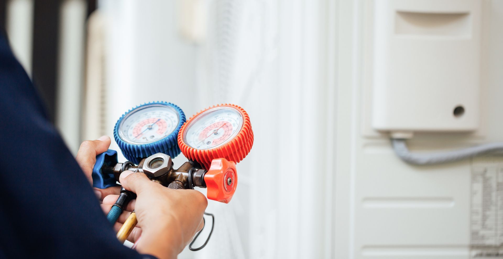 Hands holding AC manifold gauges near an outdoor air conditioning unit.