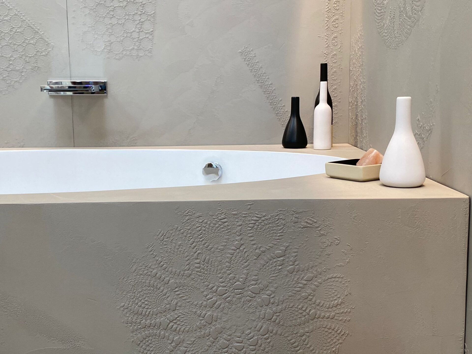 Bagno minimalista con vasca bianca, pareti beige con finitura materica e vasi bianchi e neri sul ripiano.
