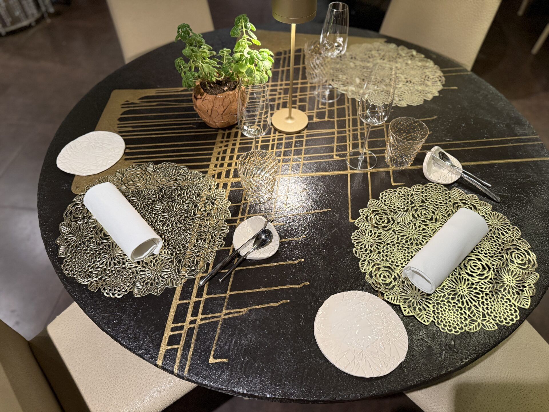 Tavolo da pranzo rotondo con tovaglia nera e oro, tovagliette intrecciate, tovaglioli bianchi, candele e una piccola pianta in vaso.