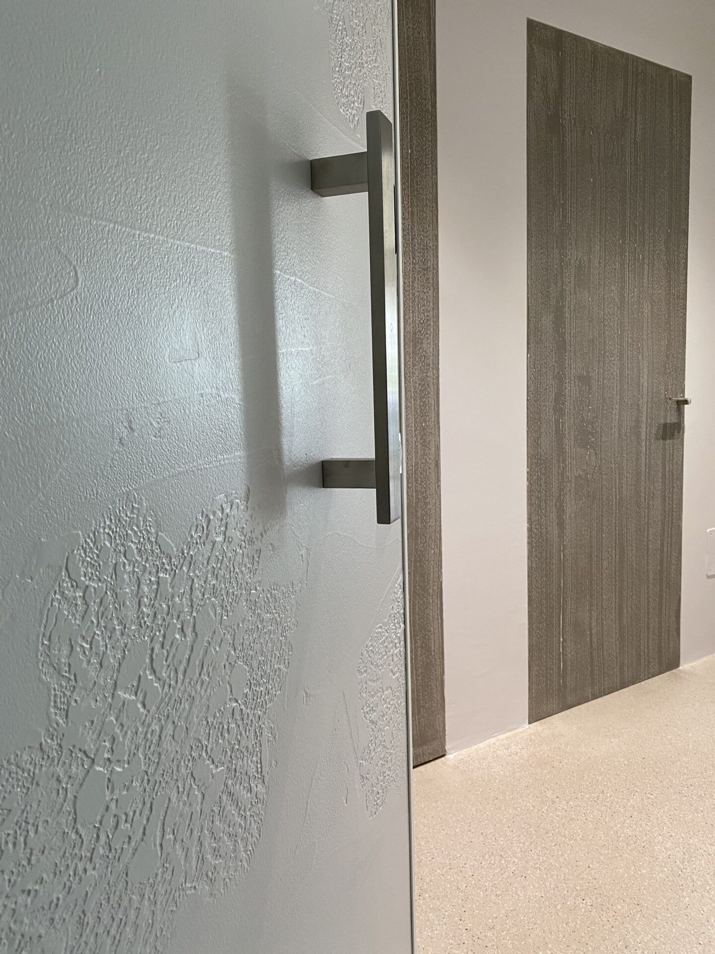 Corridoio con parete bianca texturizzata, maniglia in metallo e porta di legno chiusa.