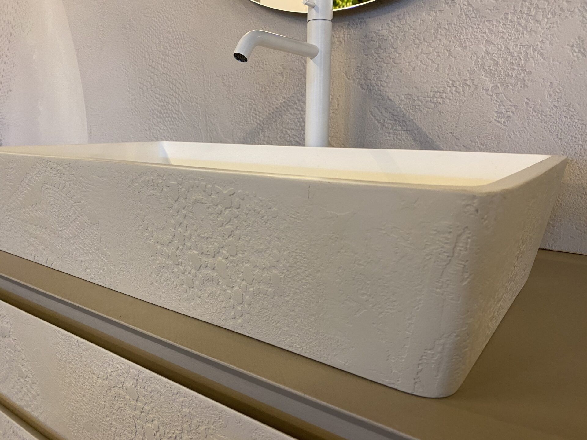 Lavabo rettangolare moderno di colore bianco con rubinetto cromato, posizionato su una parete chiara con finitura materica.