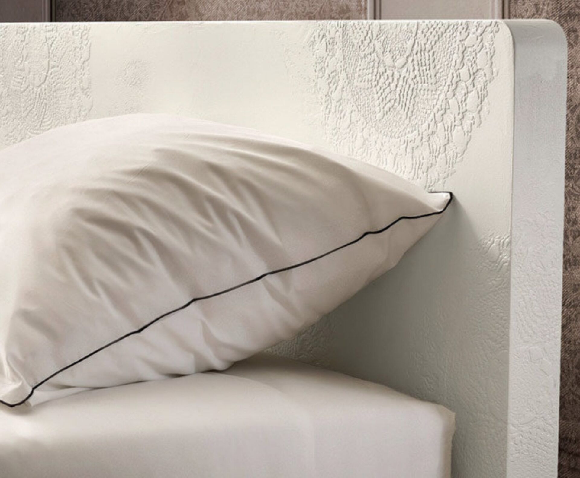 Un cuscino bianco su un letto, contro una parete color crema con una texture particolare, e una testiera imbottita grigia.