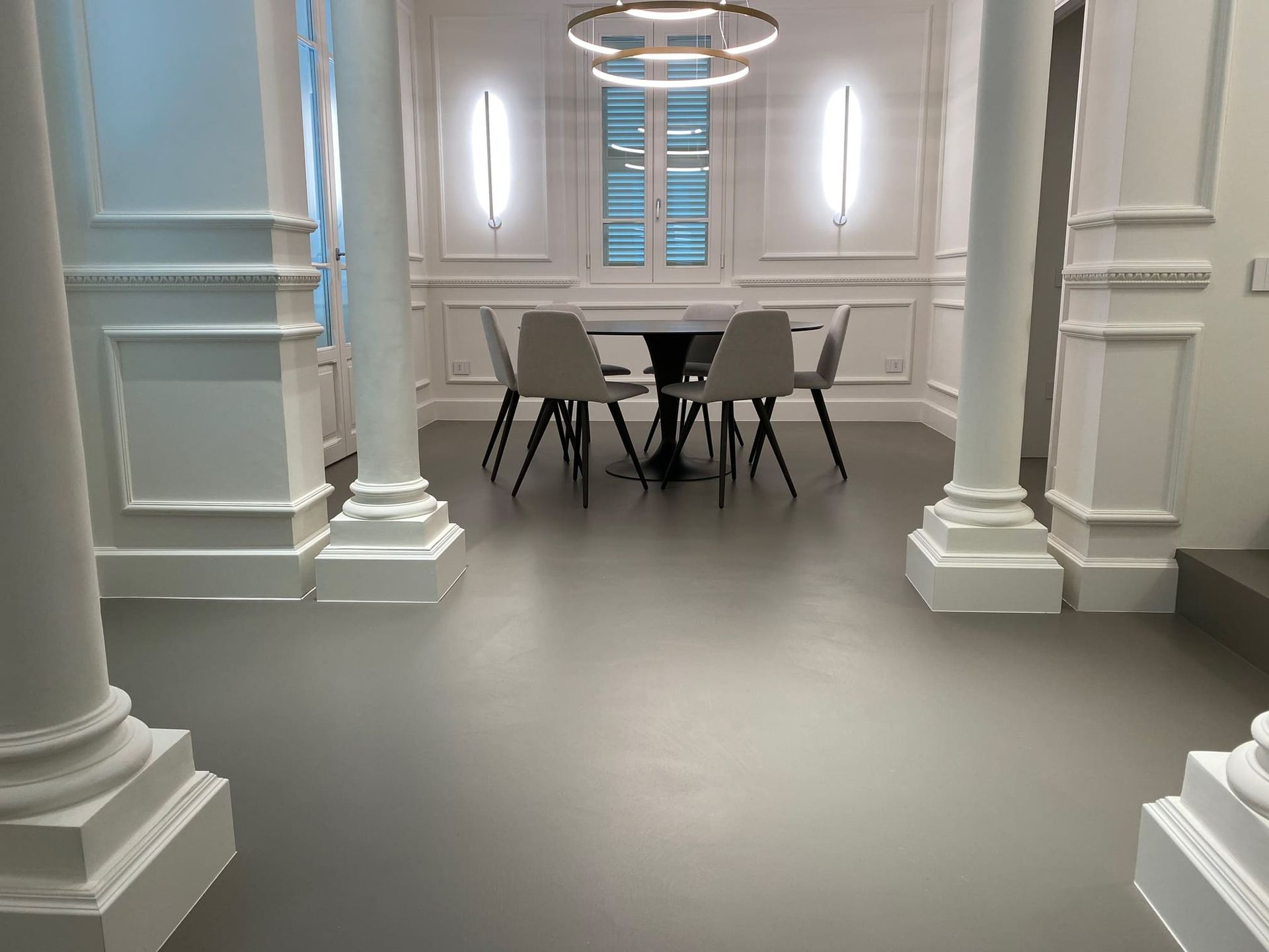 Elegante sala da pranzo bianca con tavolo rotondo e sedie tra grandi colonne e applique a parete illuminate.