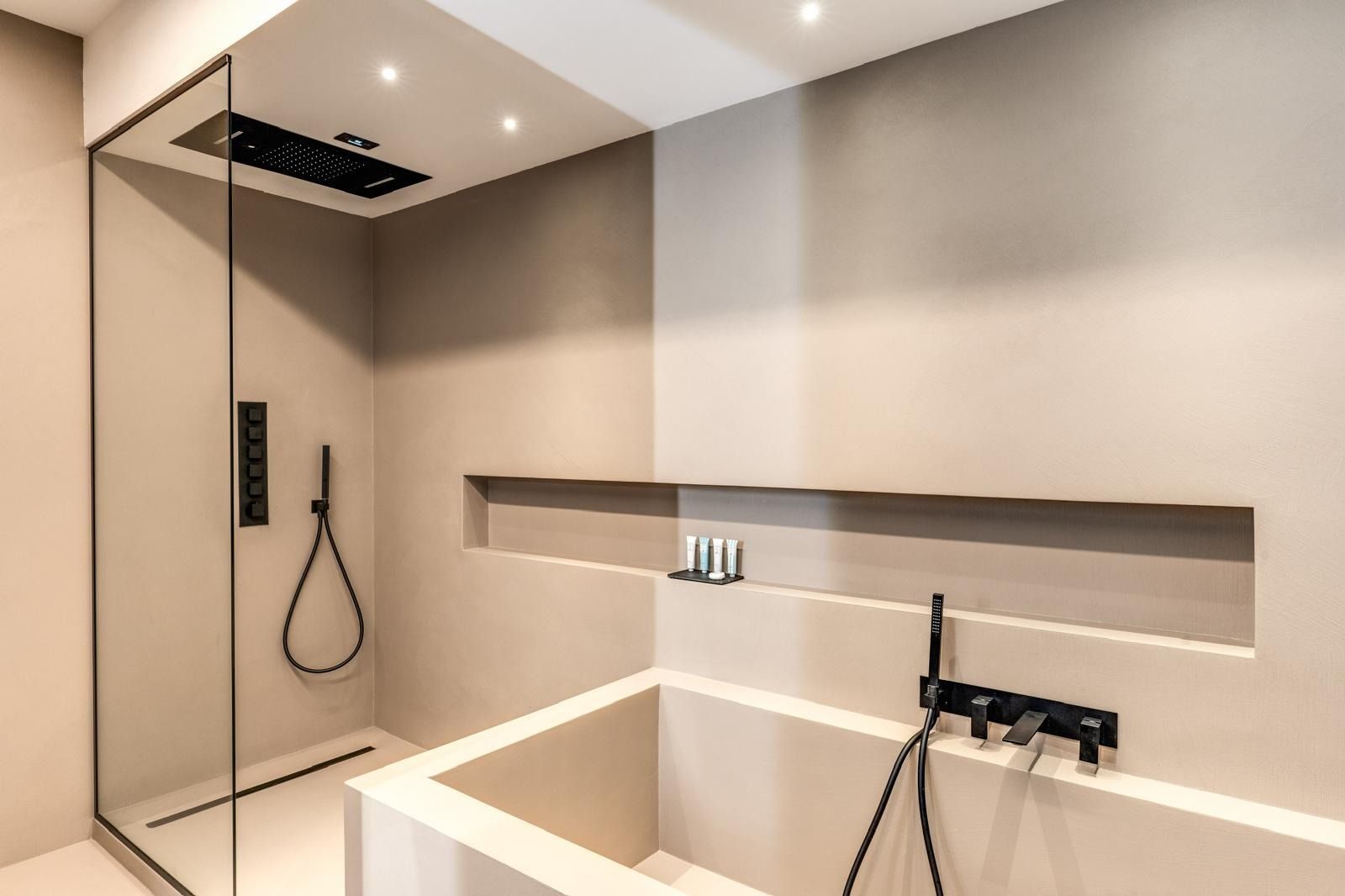 Bagno minimalista sui toni del beige con cabina doccia, rubinetteria nera e mensola a muro.