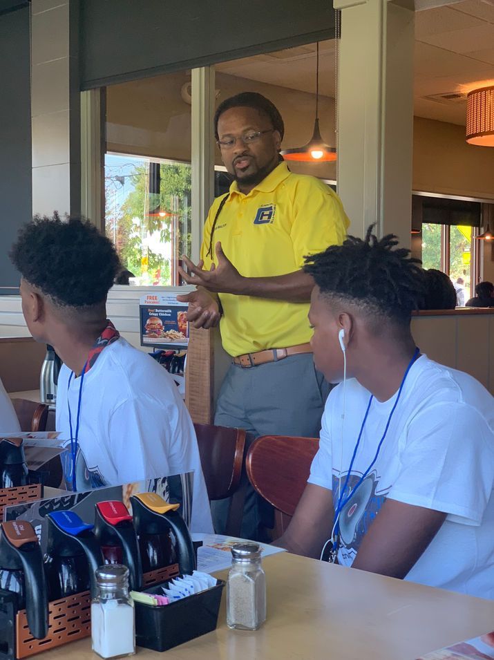 A man in a yellow collared shirt speaks to two individuals wearing white shirts, seated at a table in a restaurant.