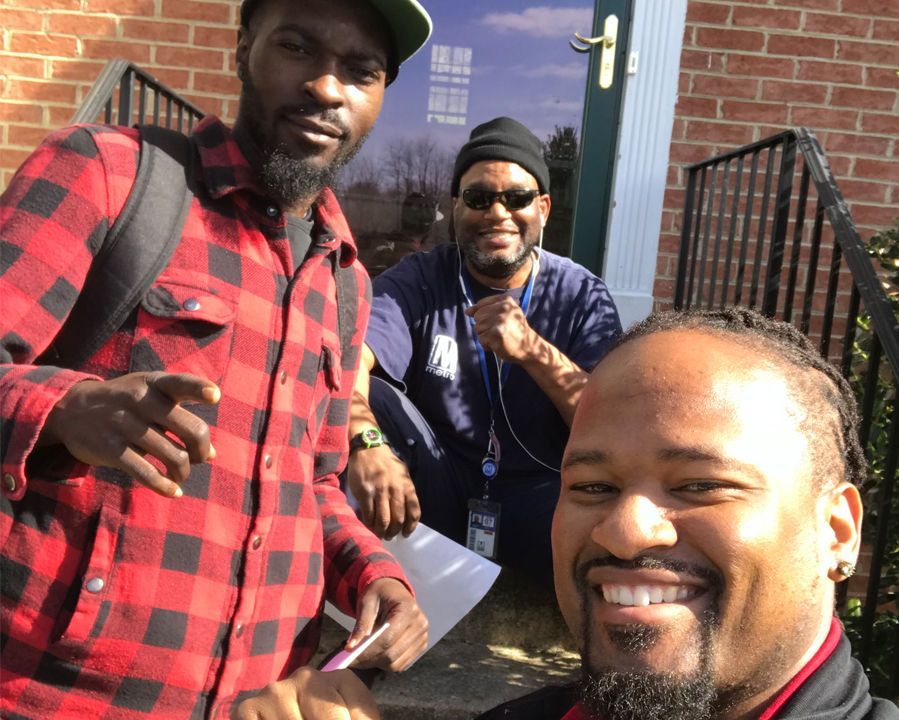 Three people smiling outside a brick building, with one person in the foreground taking a selfie.