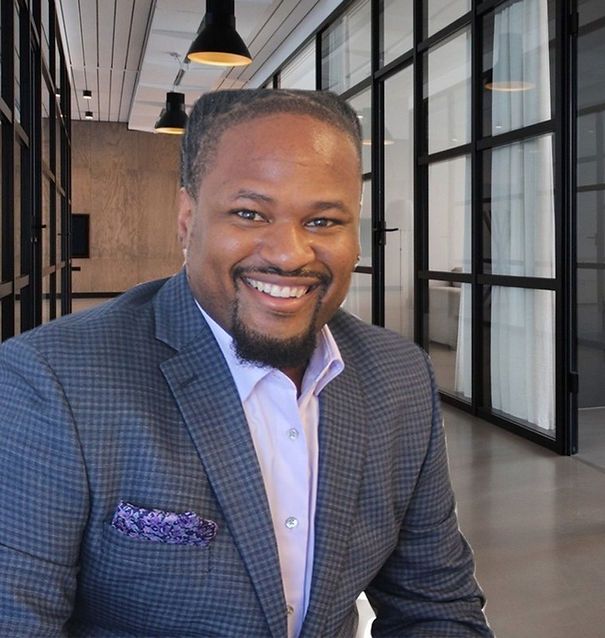 Man in a blazer smiles, in a modern office with glass walls.