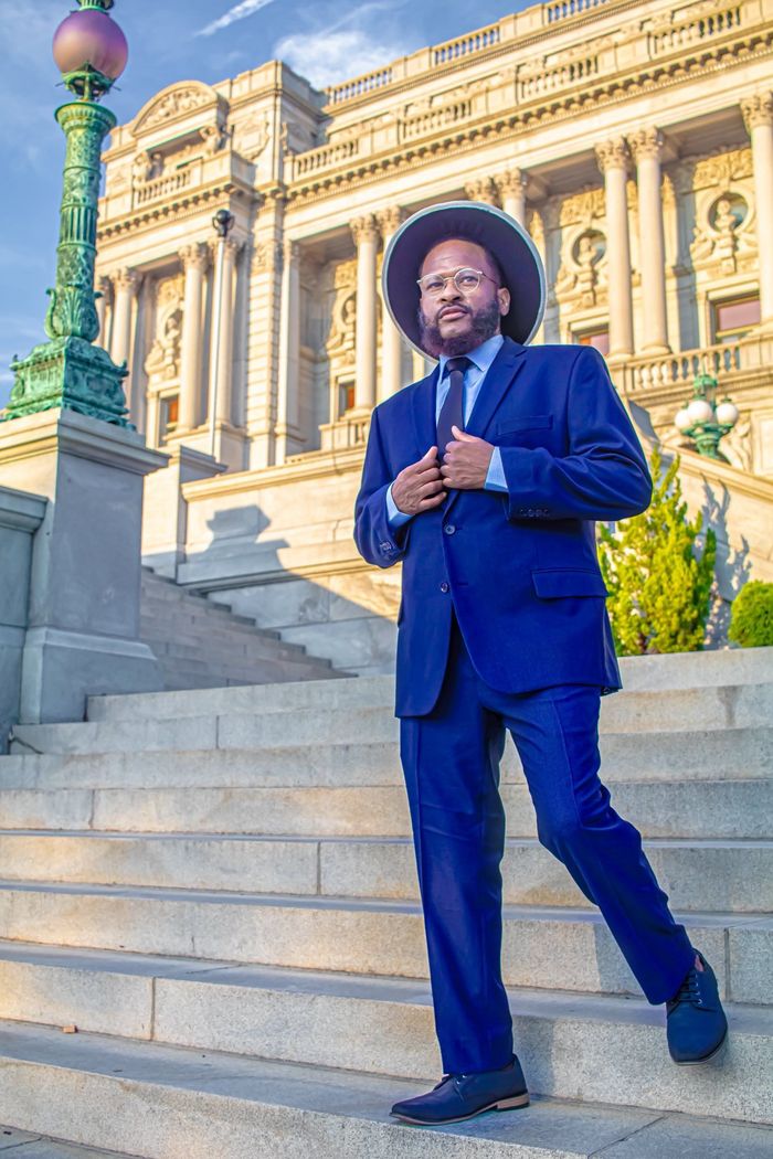 Man in a blue suit and hat poses on steps in front of a classical building.