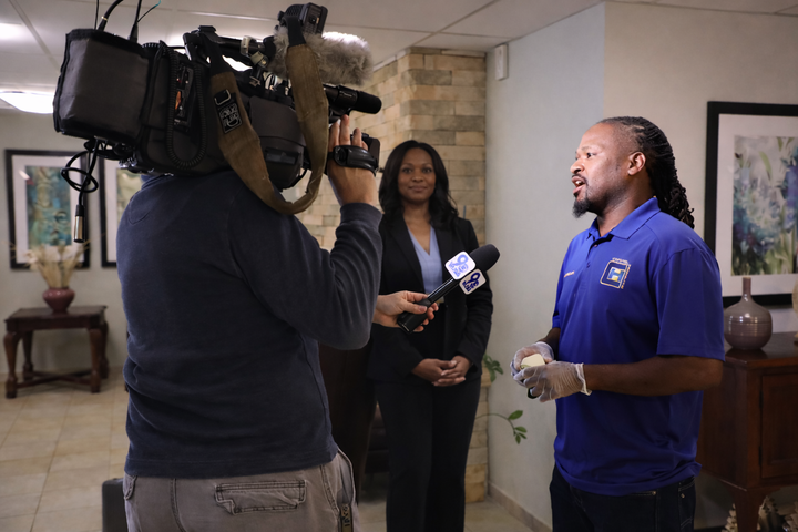 A videographer records an interview of a man in a blue polo shirt held by a person in a black suit in a lobby.