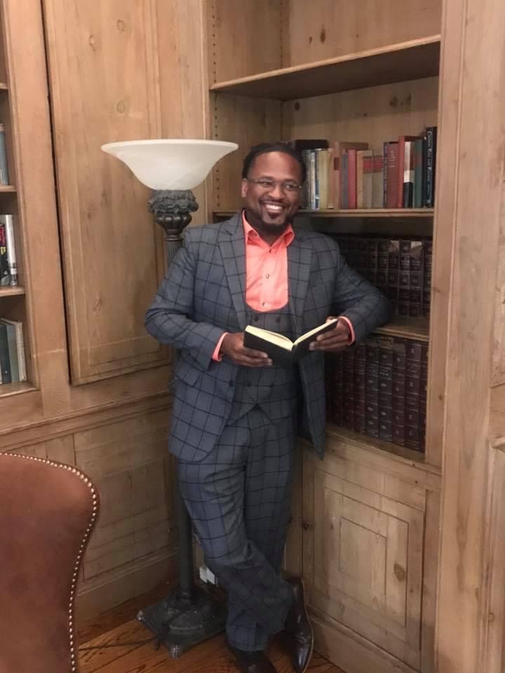Four people smile closely together in an outdoor setting with a paved path, trees, and a fence in the background.