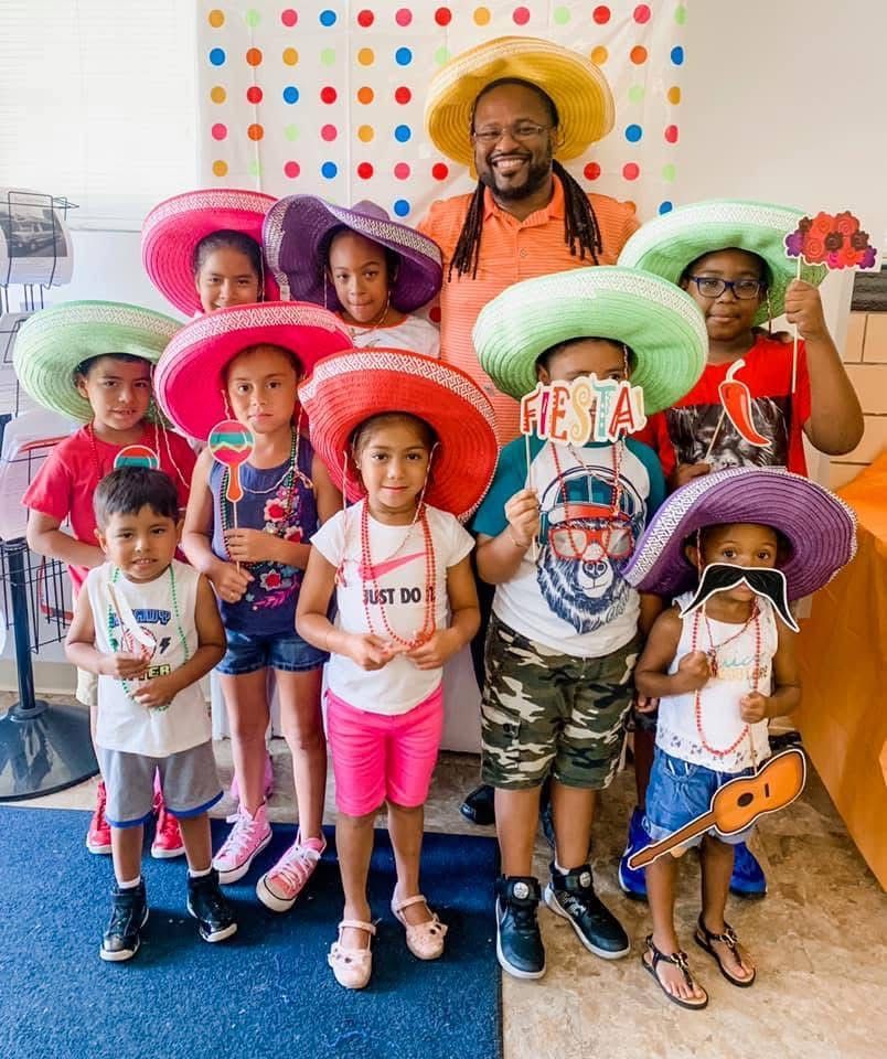 A group of children and an adult wearing colorful sombreros and holding festive props in a polka-dot decorated room.