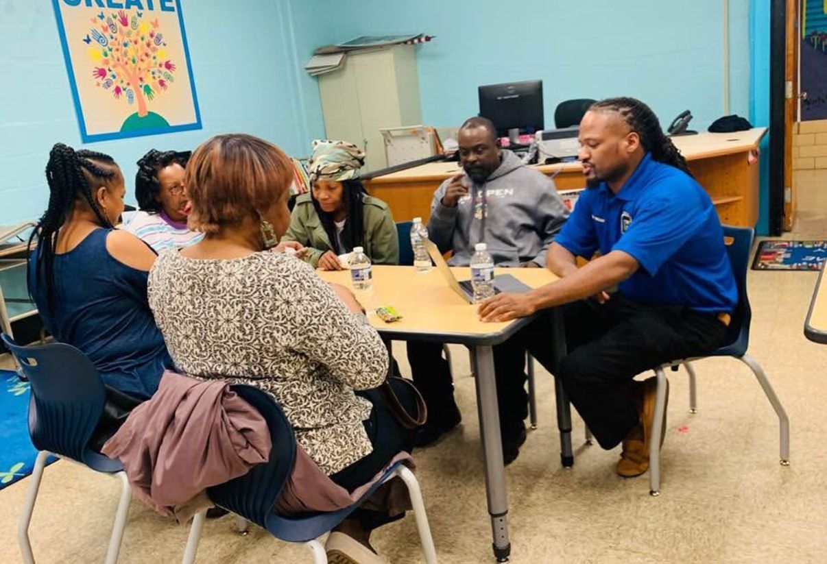 A group of six people sit around a table in a classroom discussing work, with laptops open in front of them.