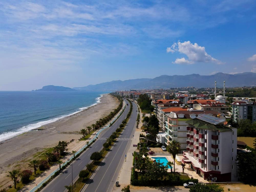 Vista aérea de uma estrada costeira junto a uma praia, ladeada por palmeiras, hotéis com piscinas e uma mesquita ao longe.
