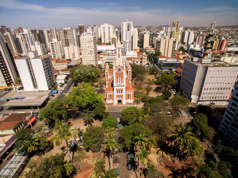 Vista aérea de uma praça da cidade com uma igreja histórica proeminente, de cor clara, rodeada por árvores verdejantes.