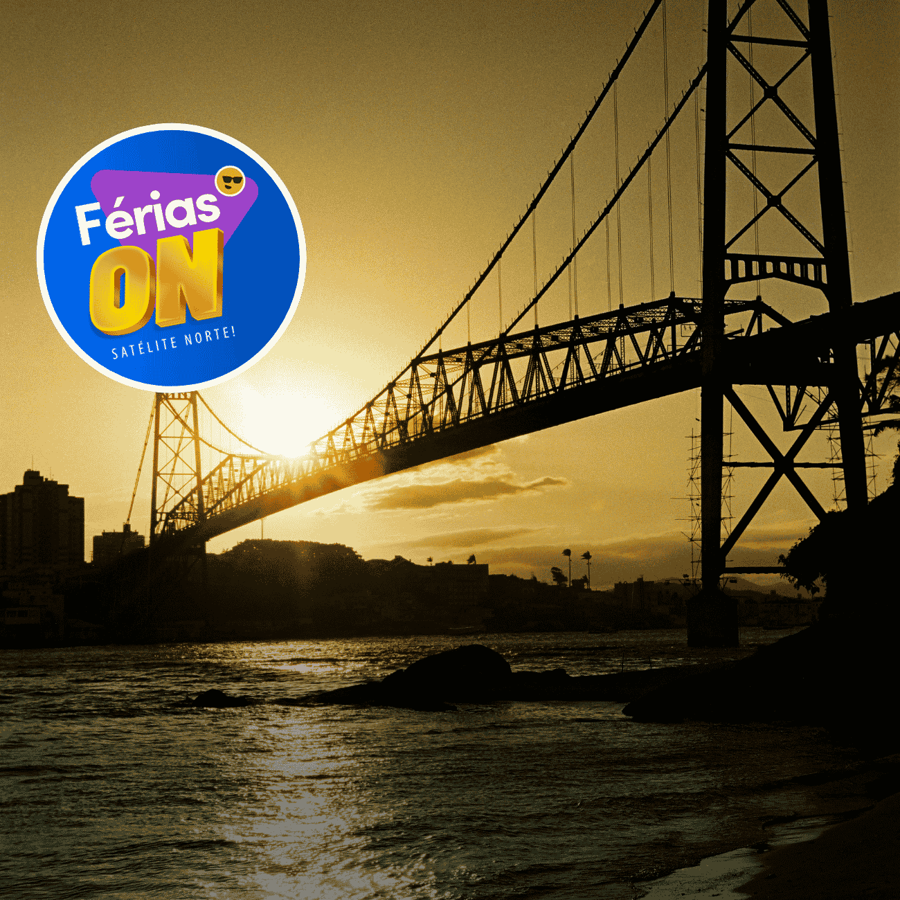 A imagem mostra uma grande ponte suspensa atravessando um corpo d’água ao pôr do sol. A estrutura aparece em silhueta escura, destacando seus cabos e torres metálicas contra um céu dourado iluminado pelo sol baixo no horizonte. A luz reflete na superfície da água, criando um caminho brilhante que se estende em direção à margem arenosa visível no canto inferior direito.