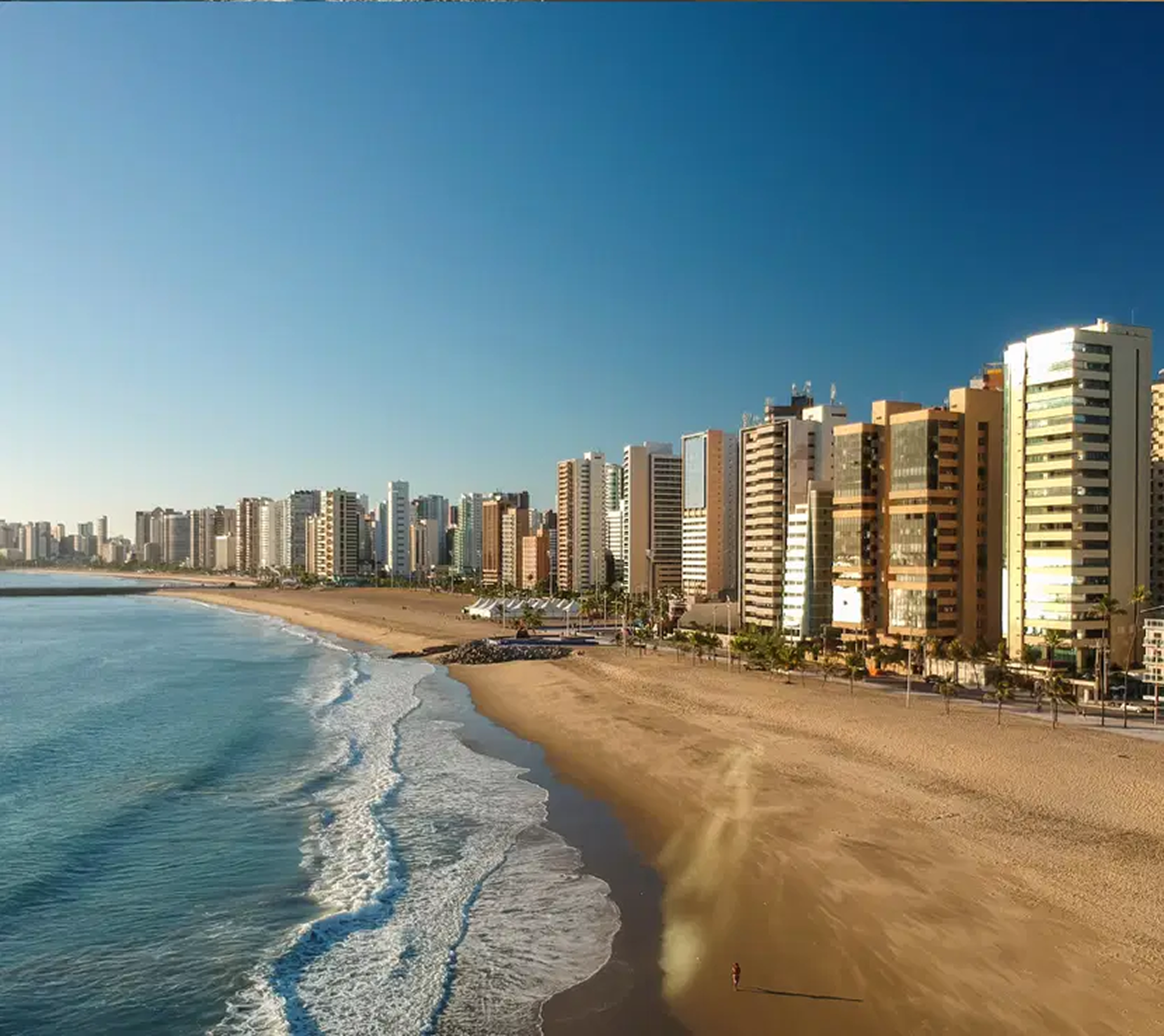 Uma praia de areia estende-se ao longo de uma costa com altos edifícios residenciais sob um céu azul claro e brilhante.