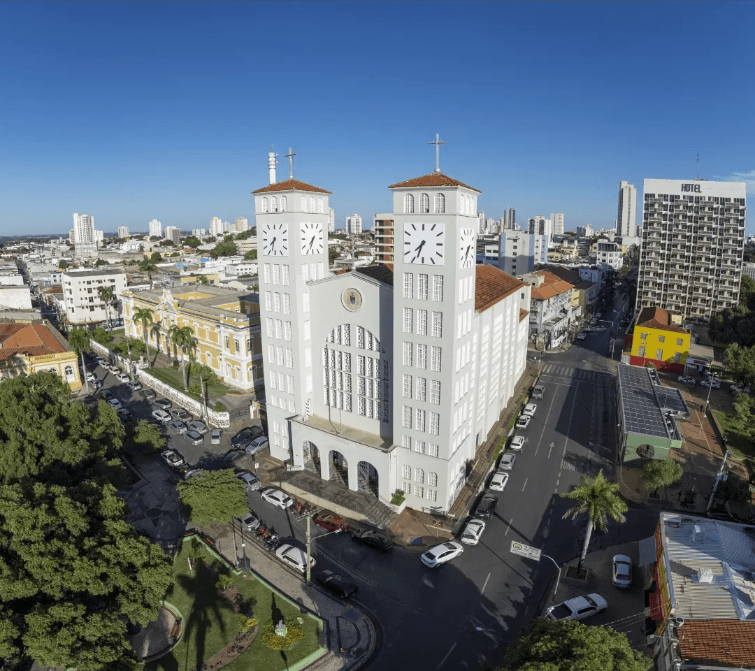 Vista aérea de uma igreja branca com duas torres em uma cidade, com prédios, árvores e carros estacionados ao redor.