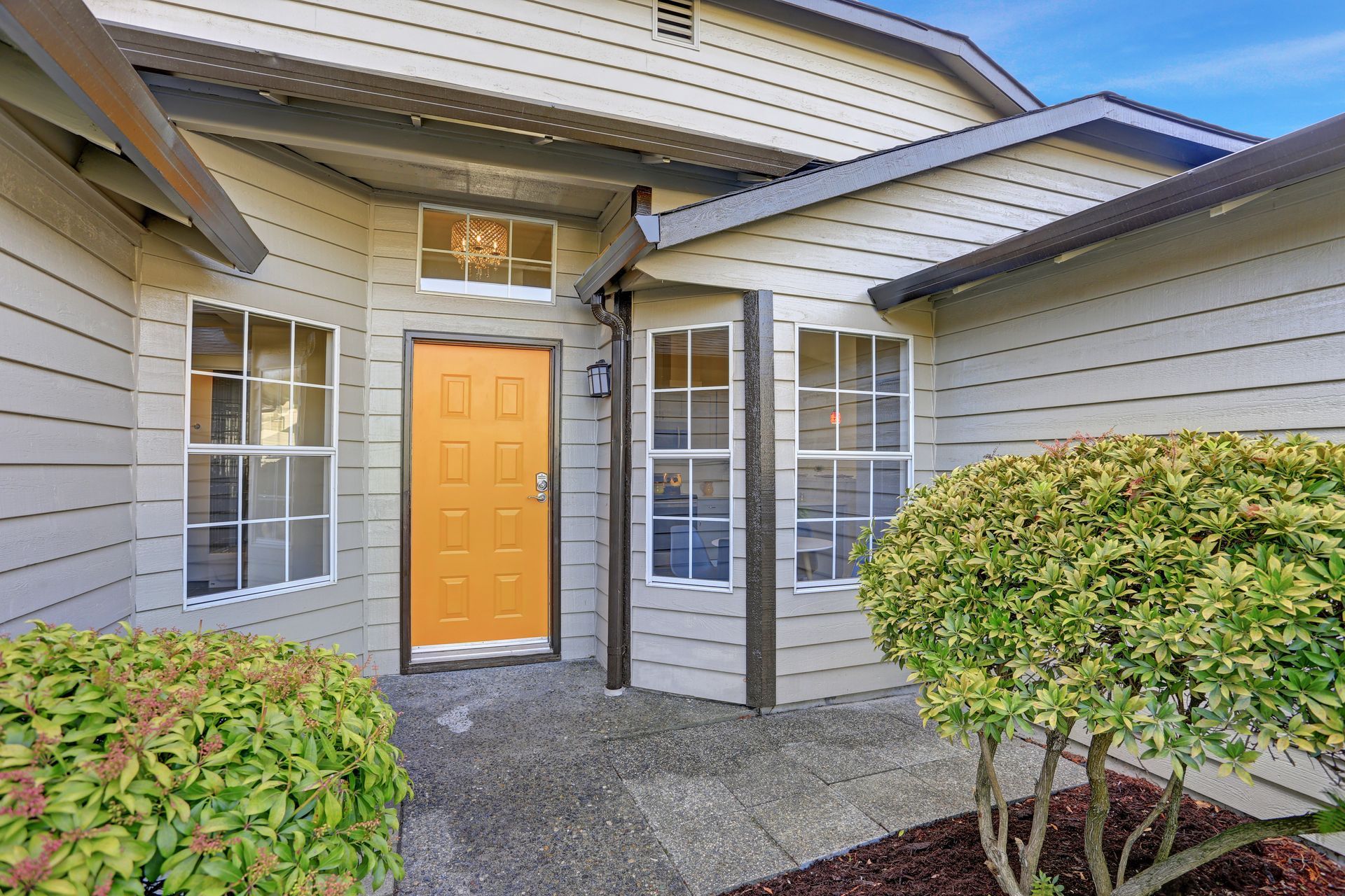 Exterior of a house with a tan exterior, orange door, and two windows. A bush sits in front.