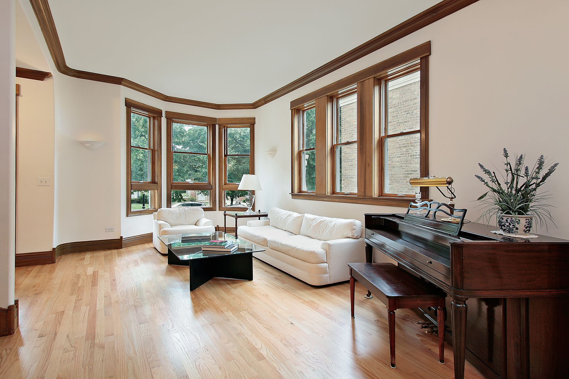 Living room with hardwood floors, white walls, and wooden trim. Furnished with white sofa, piano, and windows.