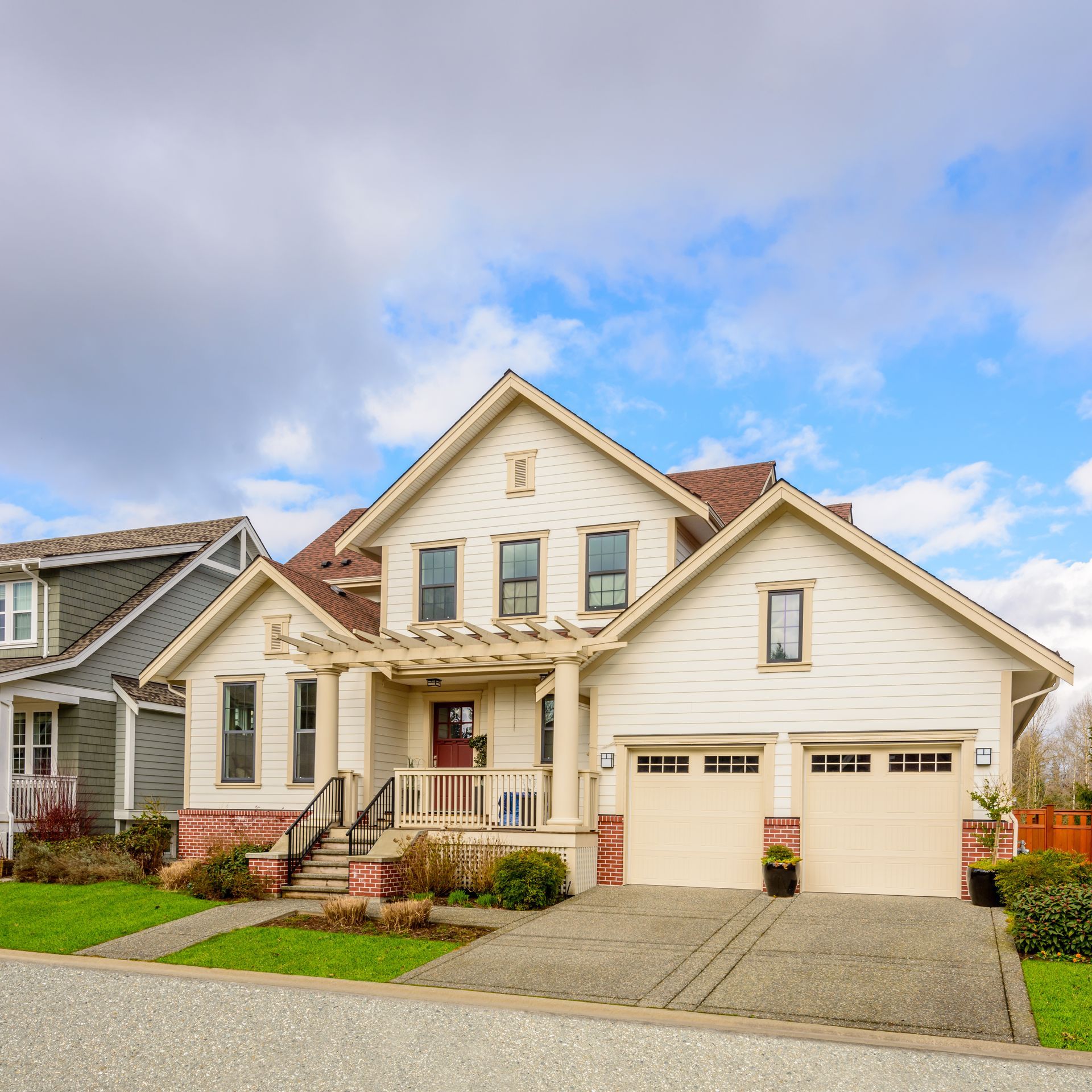 Beige two-story house with a two-car garage, red door, and pergola. Blue sky with clouds.