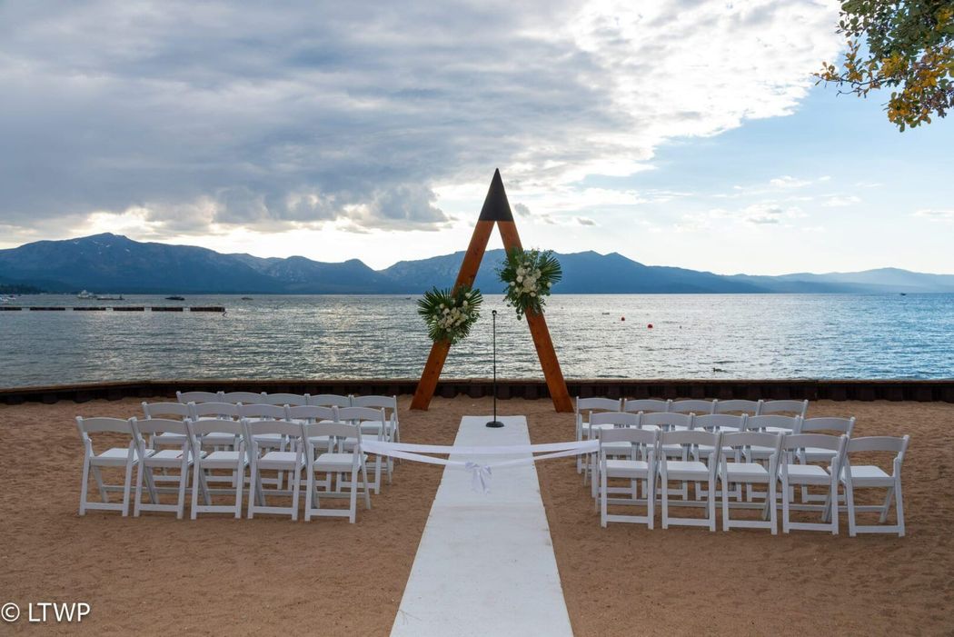 White chairs with blue flower arrangements line a grassy aisle toward a large tree and water view, set for a ceremony.