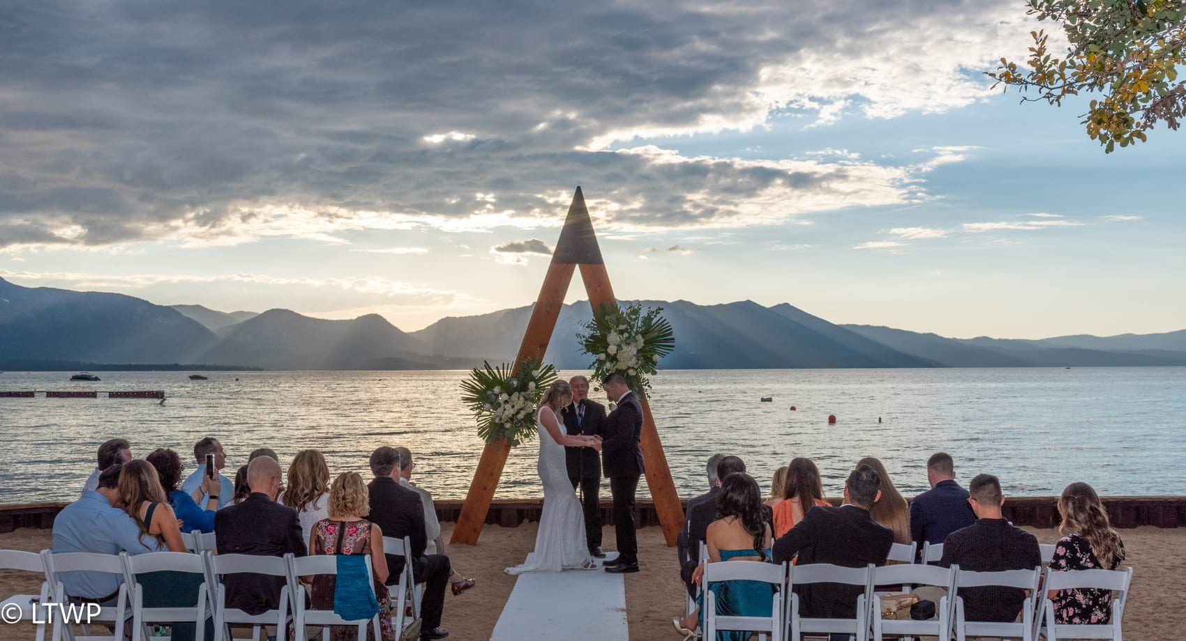 Wedding ceremony by a lake at sunset. Couple kissing under a wooden arch; guests seated on chairs.