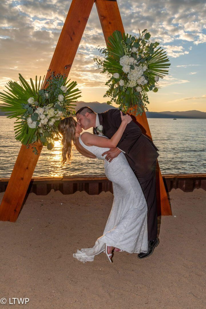 Bride and groom kissing, leaning back, during sunset, beside a wooden arch decorated with flowers, waterfront background.