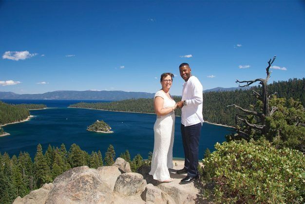 Couple holding hands, smiling, overlooking lake with island on a sunny day.