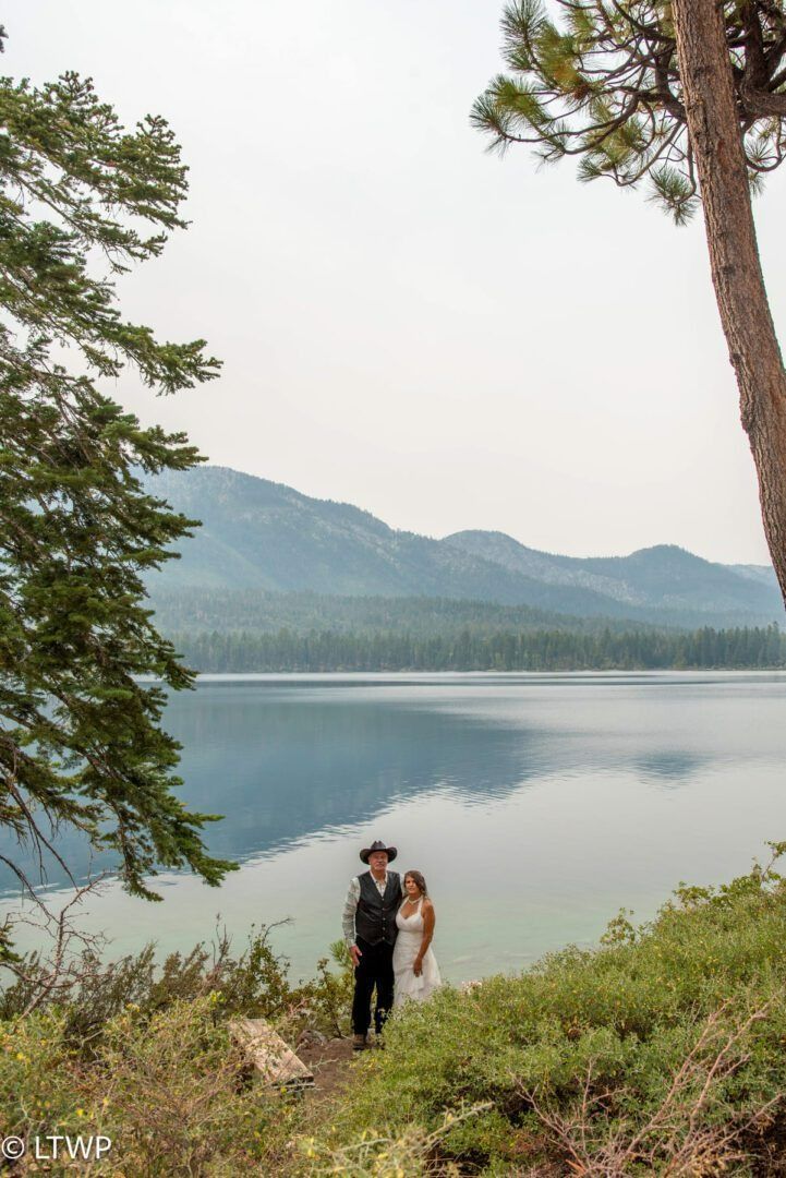 Couple standing by lake; mountains in background. Overcast sky.