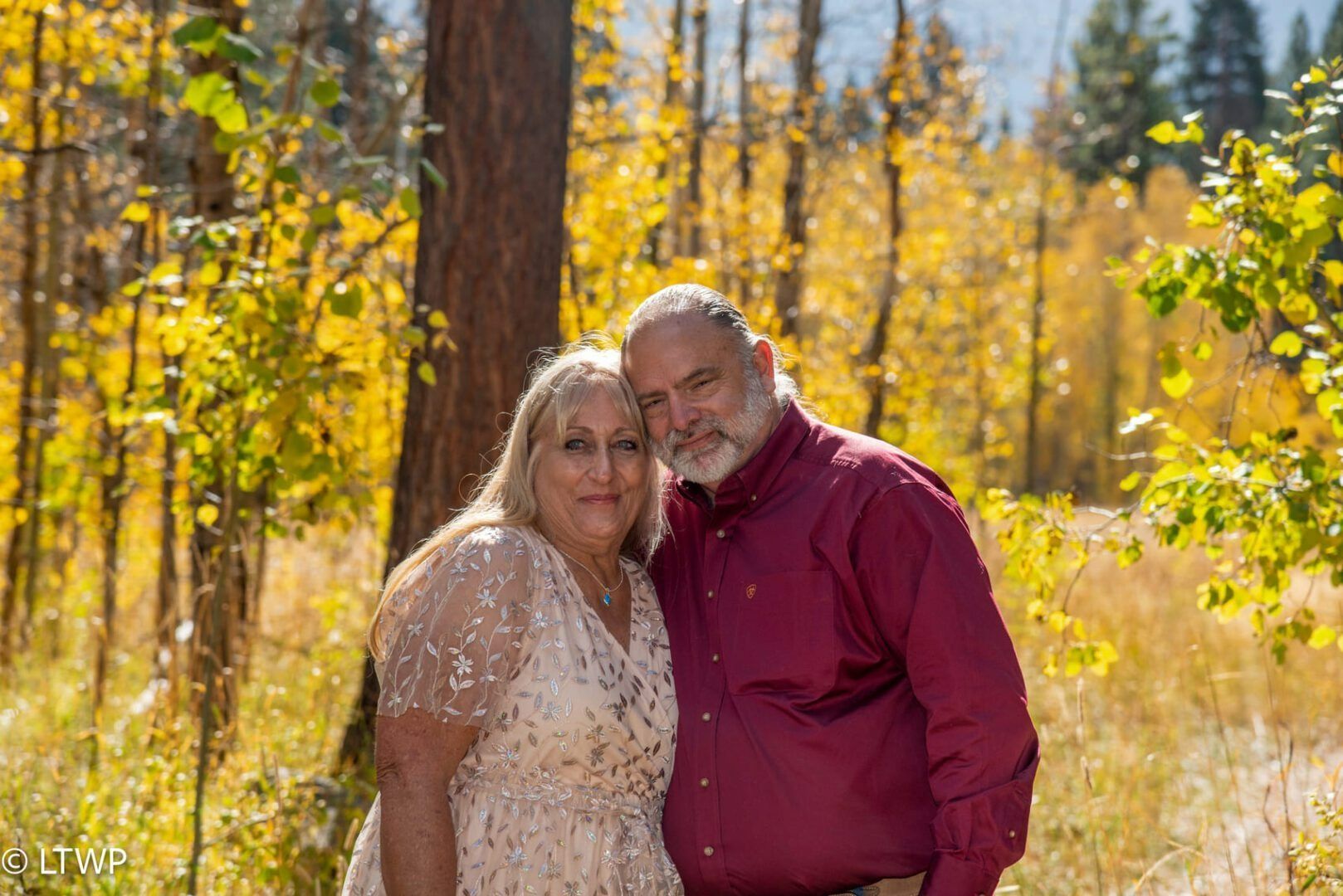 Couple embracing outdoors, surrounded by yellow foliage. Man in maroon shirt, woman in light dress.