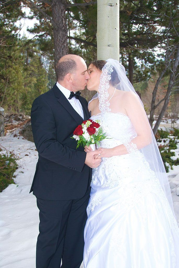 Bride and groom kissing outdoors in snow, holding bouquet. Groom in tuxedo, bride in white dress and veil.