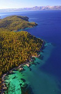 Emerald-green and deep-blue water meets a lush green, forested shoreline. Mountains are in the distance.