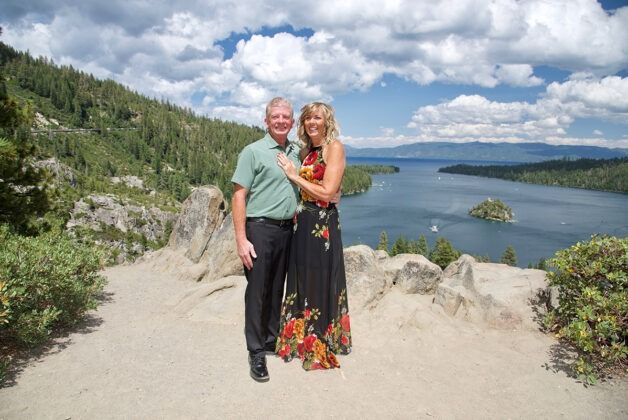 Couple poses on a scenic overlook of a lake, mountains, and blue sky.