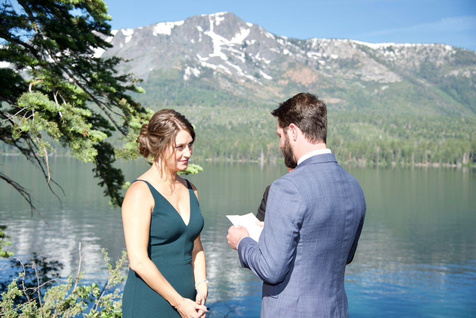 Couple at a lakeside wedding ceremony; man reading vows, woman smiling, mountain backdrop.