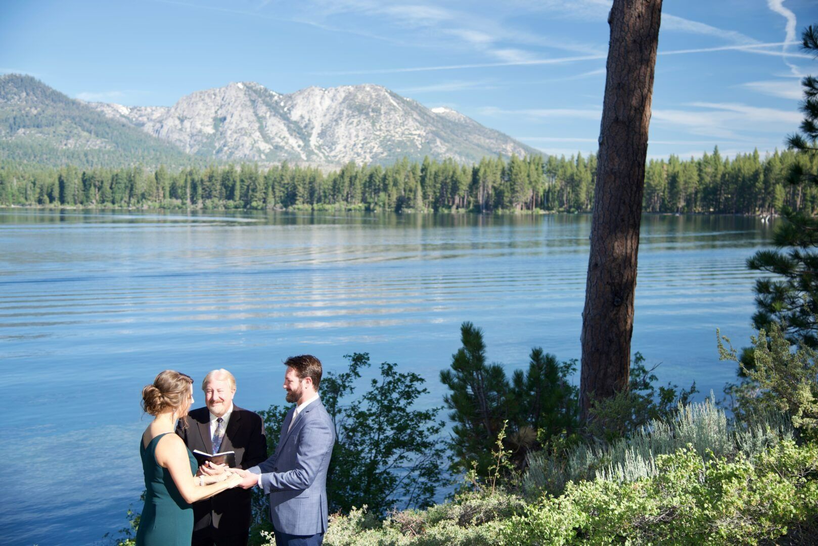 Wedding ceremony by lake with mountains in background. Couple holding hands, officiant in middle. Blue water and sky.