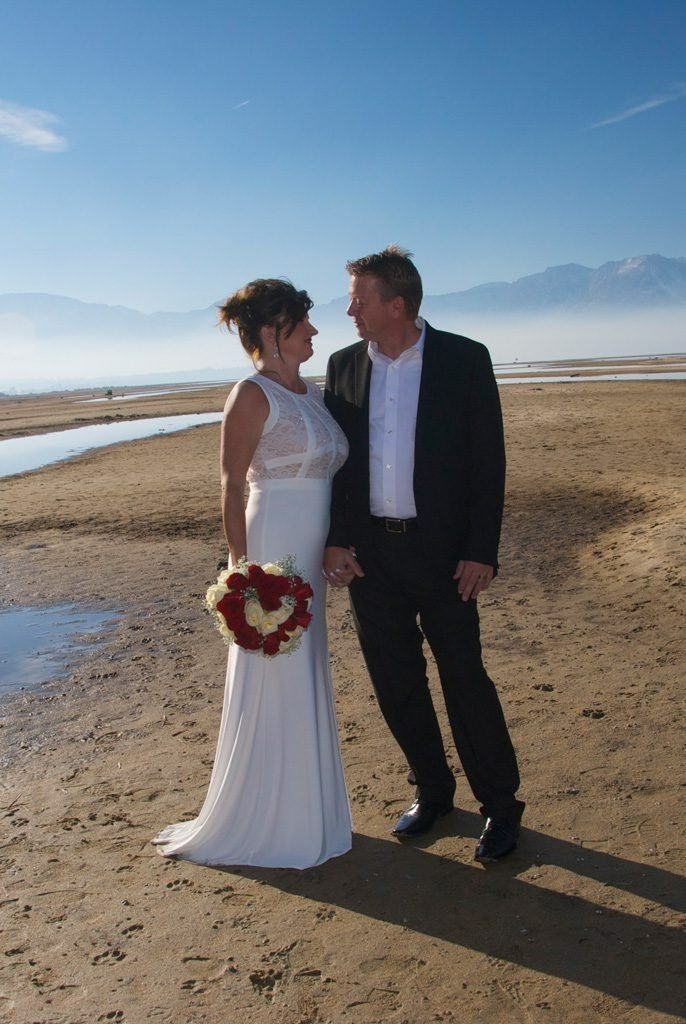 Couple in wedding attire on a beach, holding hands, looking at each other. Mountains and blue sky in the background.