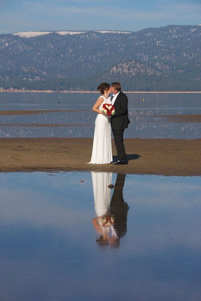 Bride and groom kissing on beach, holding red flowers. Water reflects the couple and mountain backdrop.