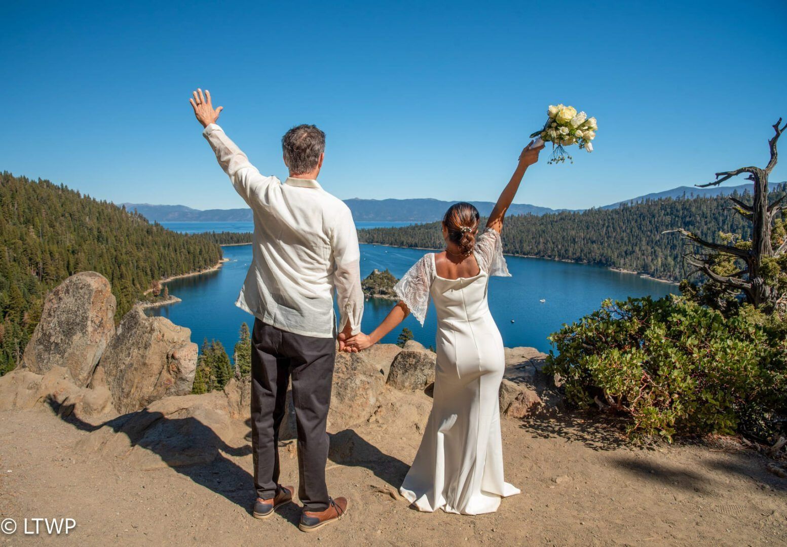 Couple on cliff overlooking lake, arms raised in celebration, bride holding bouquet.