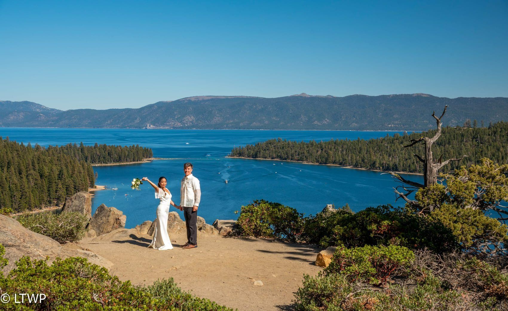 Couple on a cliff overlooking a blue lake, holding flowers, surrounded by green trees and mountains.