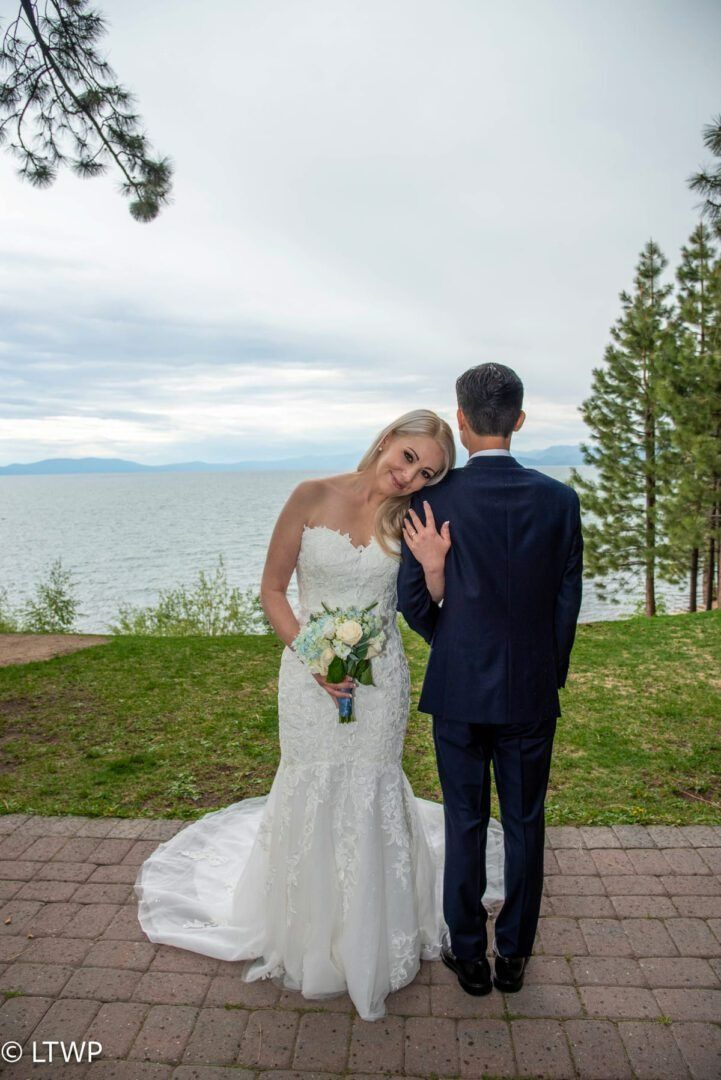 Bride in a white lace dress leans on the groom, both facing away, by the water.
