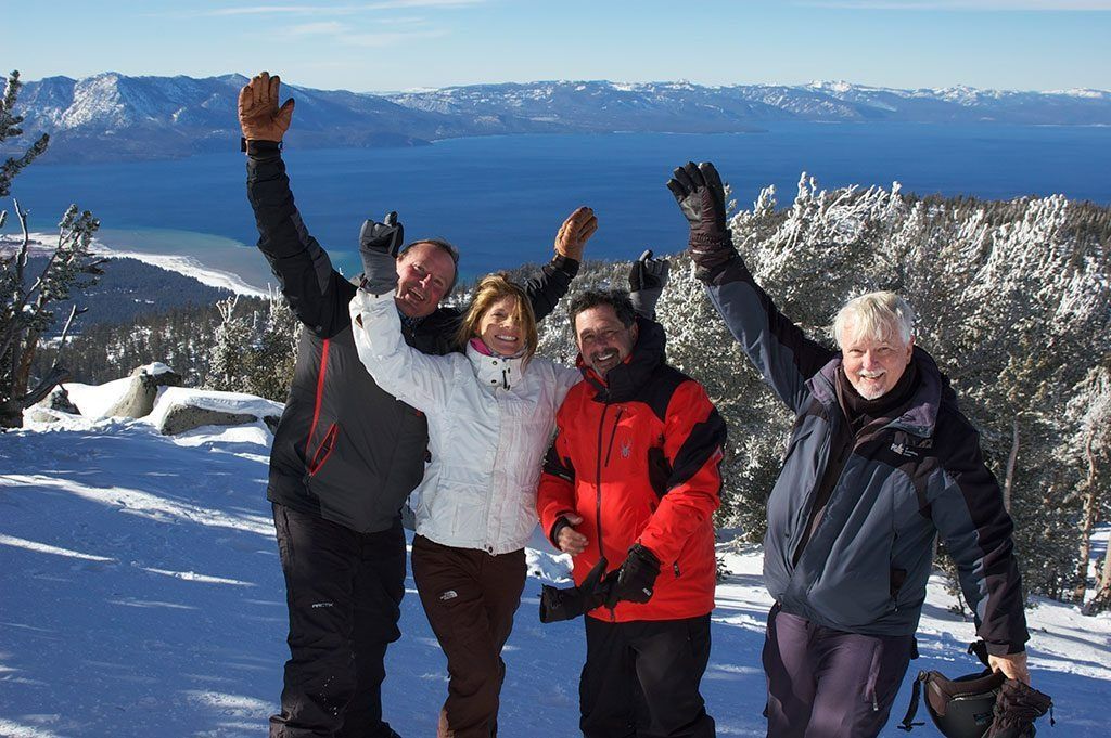 Four people in winter gear raise arms joyfully on a snowy mountain, blue lake in background.