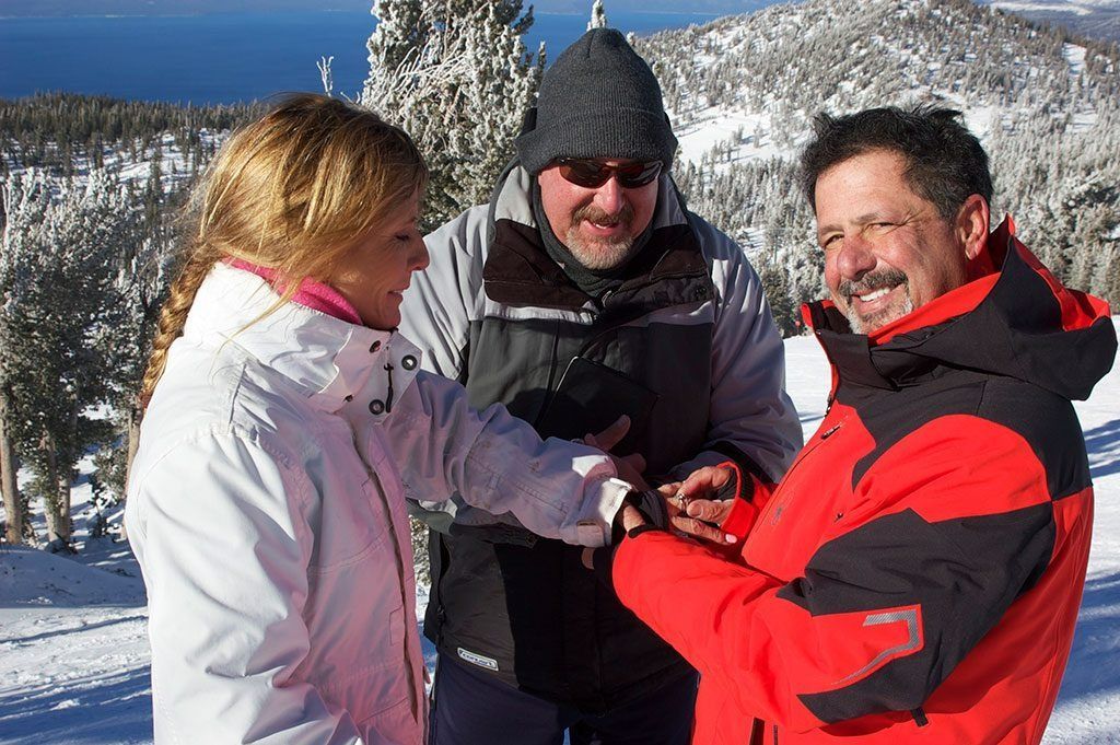 People in winter gear holding hands on a snowy mountain with a view of a blue body of water.