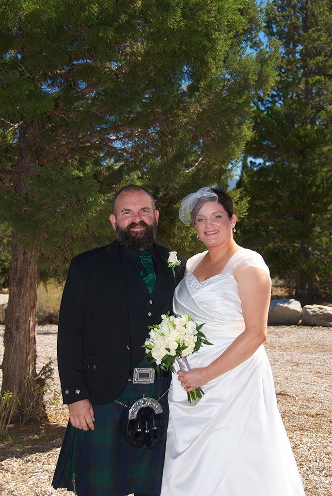 A couple in wedding attire poses outdoors; man in kilt, woman in white dress, holding flowers.